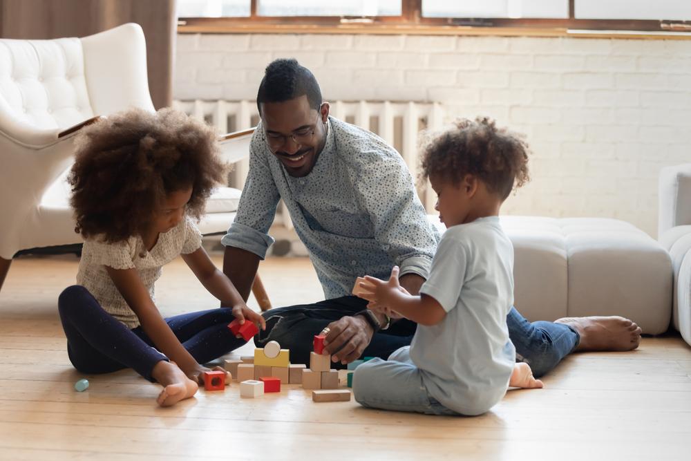 Interested black dad engaged in building wooden brick towers with a school-age girl and little boy at home
