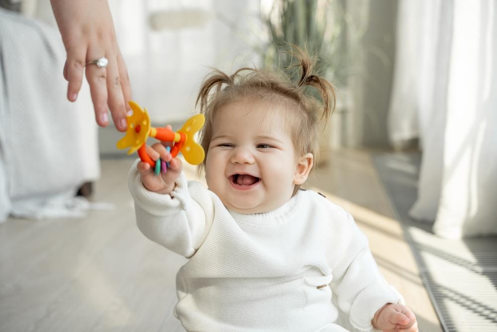 Mom giving to smiling baby favorite toy rattle teether. Child toothache teething, dentition, growing teeth. Care of children, colorful games, Parent care of kids.