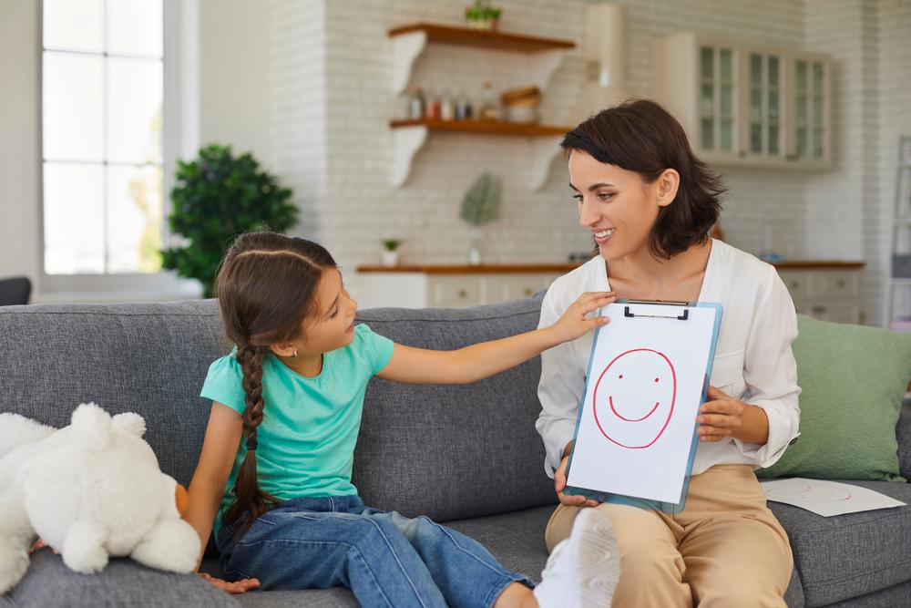 Smiling assertive psychologist with picture of happy emoji talking about emotions with 8 - 10-year-old girl.