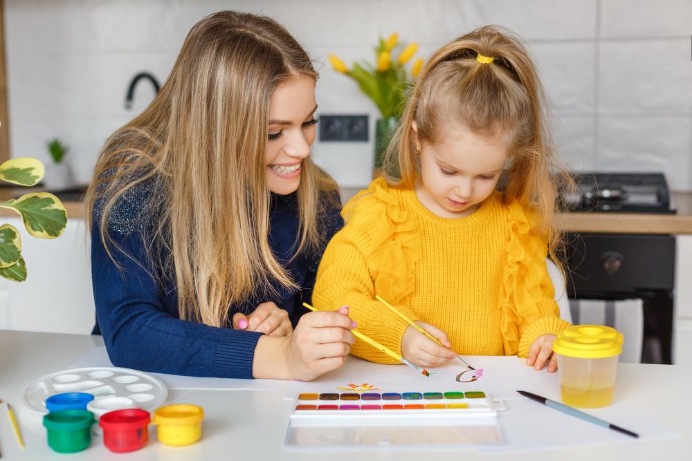Mother and daughter painting at home. Cute little kid in yellow sweater having fun with parent and paints.