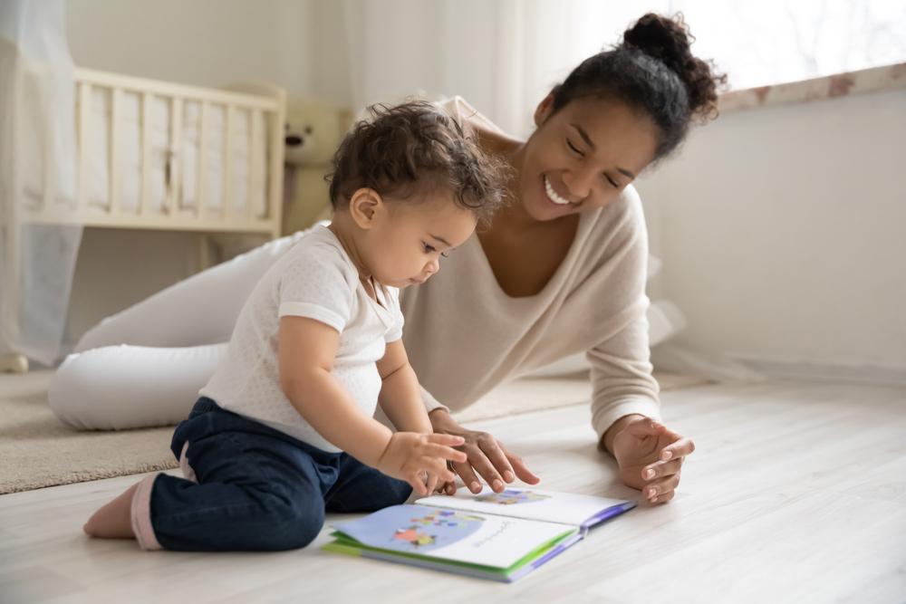 Smiling young African American mother sits on warm floor play with little infant toddler child.