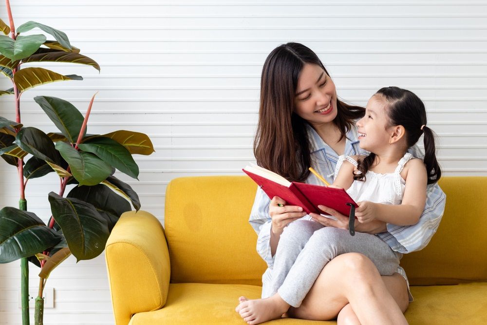 Mom and daughter reading and writing together on couch at home. Happy family and mother's day concept. Asian woman and Caucasian girl in pajamas playing at home.