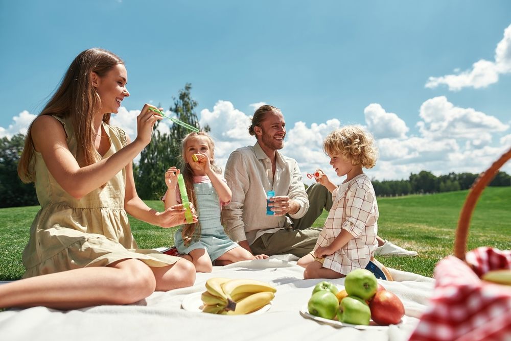 Two little kids blowing soap bubbles while spending time with their parents, family having picnic in nature on a summer day. 