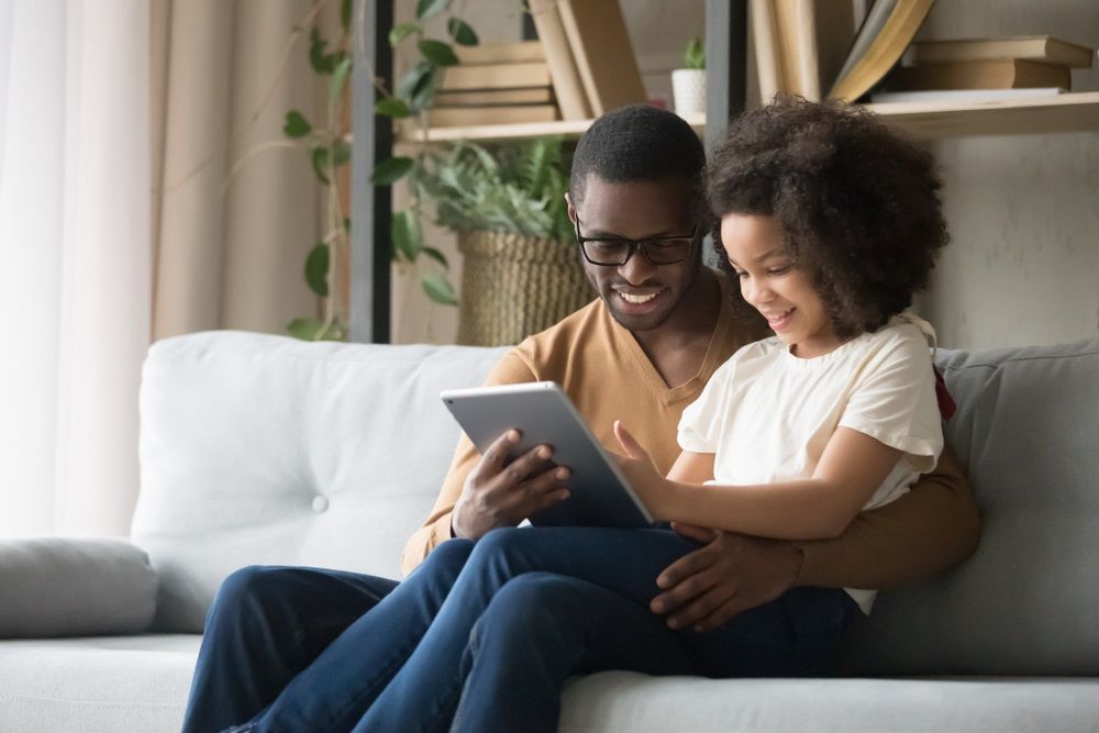 African father holds on lap daughter sitting resting on sofa in living room using digital tablet holding mobile computer watch cartoons educational online program, buying shopping via internet concept